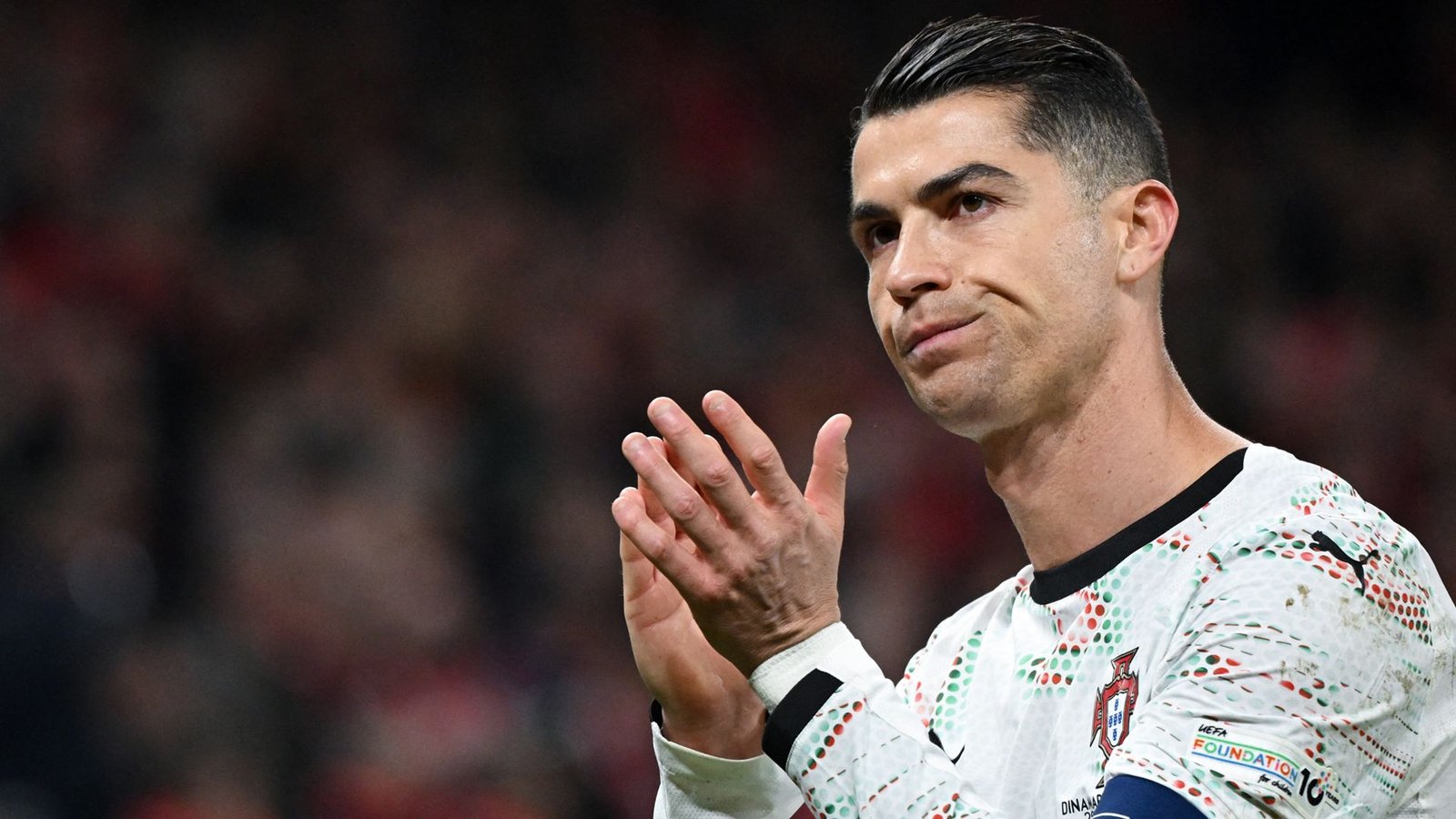 Cristiano Ronaldo applaudisseert voor fans in een witte Portugal-outfit met kleurrijke ontwerpen, met een gemengde uitdrukking tijdens een wedstrijd.