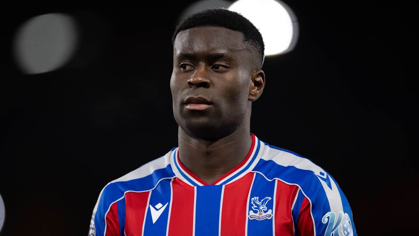 Marc Guehi in Crystal Palace's red and blue kit, displays a focused expression during a match, with blurred stadium lights behind.