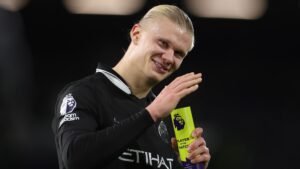 Erling Haaland sourit et salue en tenant le trophée de 'Joueur du Match', portant le maillot noir de Manchester City.