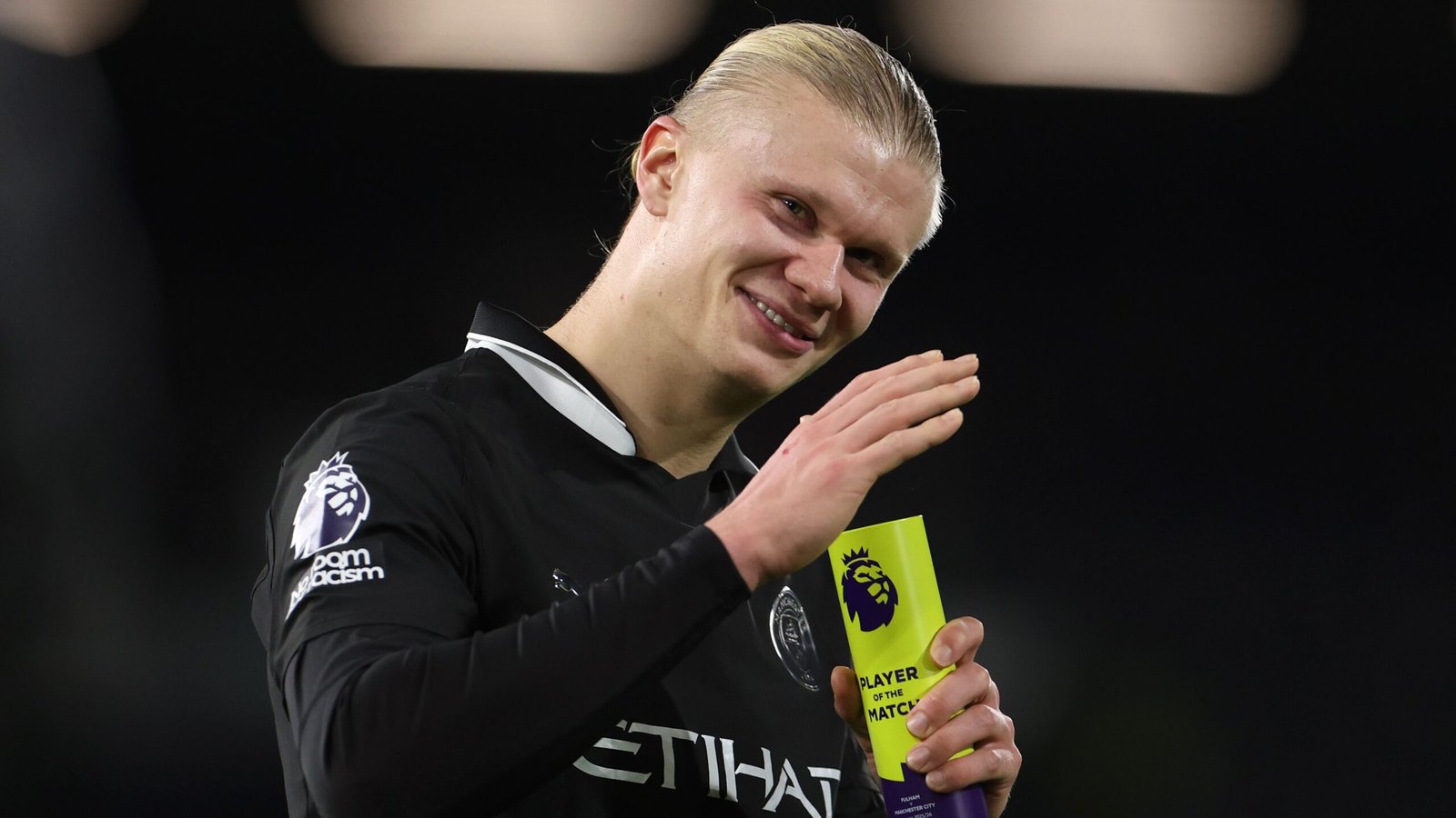 Erling Haaland sourit et salue en tenant le trophée de 'Joueur du Match', portant le maillot noir de Manchester City.
