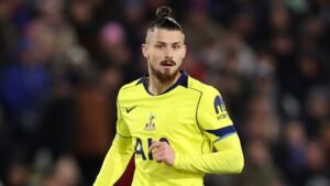 Radu Drăgușin in Spurs' felgele tenue, toont een geconcentreerde uitdrukking tijdens een wedstrijd, tegen een wazige menigte op de achtergrond.