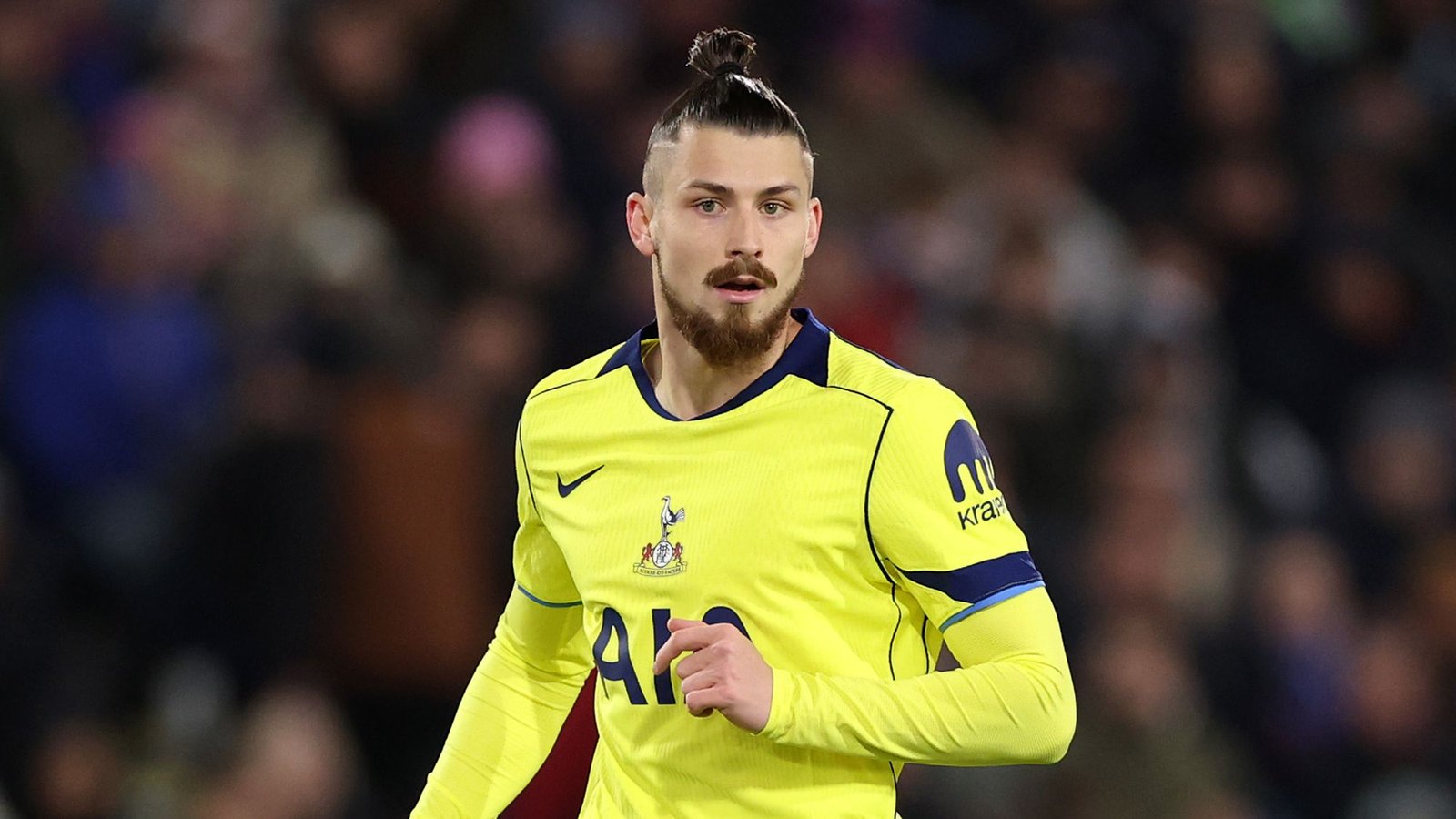 Radu Drăgușin in Spurs' felgele tenue, toont een geconcentreerde uitdrukking tijdens een wedstrijd, tegen een wazige menigte op de achtergrond.