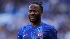 Raheem Sterling, in a blue Chelsea kit with the club logo, looks focused during a match, sweat glistening on his face.