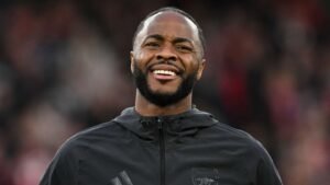 Raheem Sterling smiles while wearing a black Arsenal jacket, with a blurred crowd in the background, during matchday preparations.