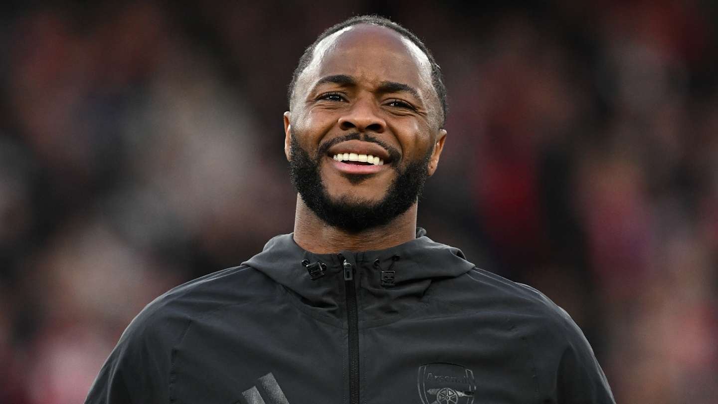 Raheem Sterling smiles while wearing a black Arsenal jacket, with a blurred crowd in the background, during matchday preparations.