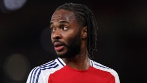 Raheem Sterling in een rood en wit voetbaltenue, die nadenkend wegkijkt tijdens een wedstrijd, met zijn kenmerkende gevlochten kapsel.
