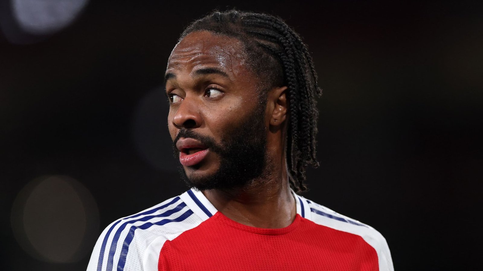 Raheem Sterling in een rood en wit voetbaltenue, die nadenkend wegkijkt tijdens een wedstrijd, met zijn kenmerkende gevlochten kapsel.
