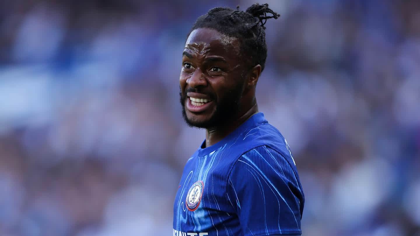 Raheem Sterling portant le maillot bleu de Chelsea, souriant avec intensité pendant un match, le stade flou en arrière-plan.