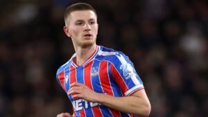 Adam Wharton dans le maillot rouge et bleu de Crystal Palace lors d'un match, faisant preuve d'une concentration et d'une détermination intenses sur le terrain.