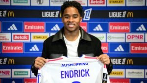 Endrick glimlacht terwijl hij zijn nieuwe Lyon-shirt vasthoudt, tegen een achtergrond van sponsoren en Ligue 1-logo's, wat zijn contractondertekening aankondigt.