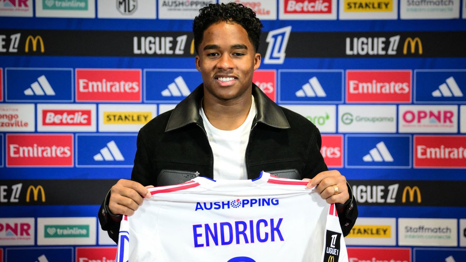 Endrick glimlacht terwijl hij zijn nieuwe Lyon-shirt vasthoudt, tegen een achtergrond van sponsoren en Ligue 1-logo's, wat zijn contractondertekening aankondigt.