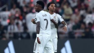 Vinícius Júnior et Rodrygo du Real Madrid, en tenues blanches, célébrant ensemble pendant un match. Des supporters en arrière-plan.