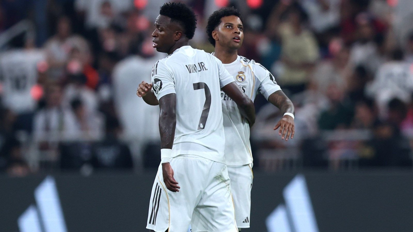 Vinícius Júnior et Rodrygo du Real Madrid, en tenues blanches, célébrant ensemble pendant un match. Des supporters en arrière-plan.