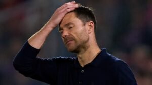 L'entraîneur déprimé se tient le front avec frustration pendant un moment tendu sur la touche, portant un polo bleu foncé.