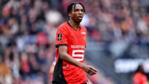 Jérémy Jacquet, joueur du Stade Rennais, portant un maillot rouge vif lors d'un match, concentré sur le jeu avec des supporters flous en arrière-plan.