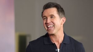 Rob McElhenney glimlacht breed, toont enthousiasme, terwijl hij in gesprek is, gekleed in een donker polo-shirt tegen een neutrale achtergrond.