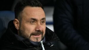 Roberto De Zerbi, vêtu d'une veste sombre, sourit en étant assis, réfléchissant à ses liens potentiels avec le poste de Manchester United.