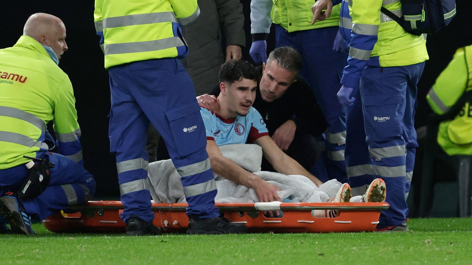 Geblesseerde voetballer die medische hulp krijgt op een brancard, omringd door paramedici in veiligheidshesjes, tijdens een wedstrijd.