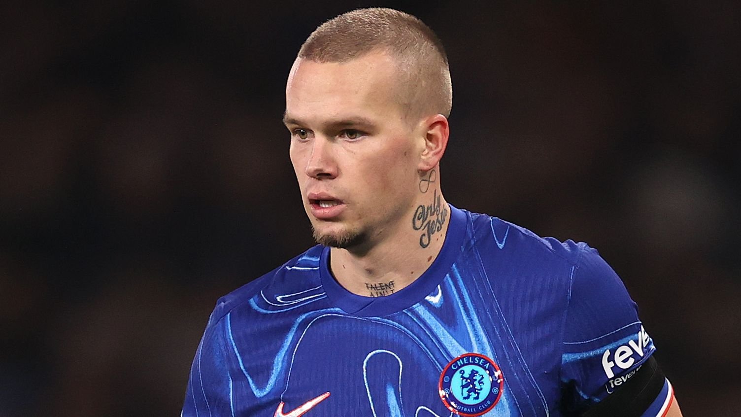 Chelsea-speler in een blauw tenue met een onderscheidend ontwerp, met een kort kapsel en zichtbare tattoos, geconcentreerd tijdens een wedstrijd.