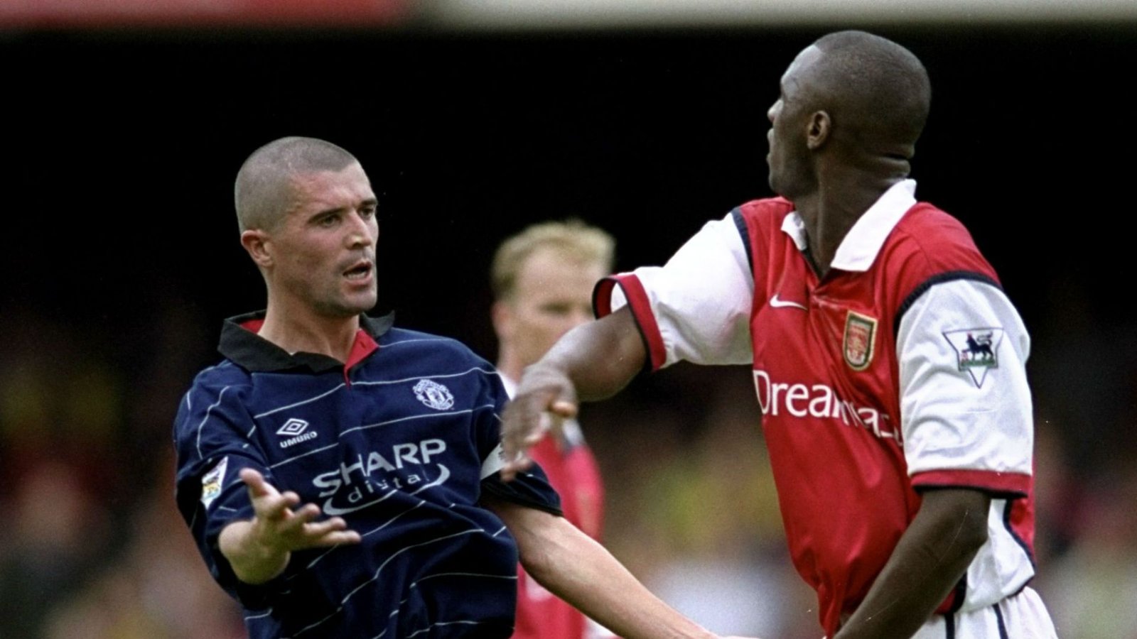 Roy Keane, in het tenue van Manchester United, gebaart levendig, terwijl Patrick Vieira, gekleed in het shirt van Arsenal, hem confronteert in een gespannen moment.