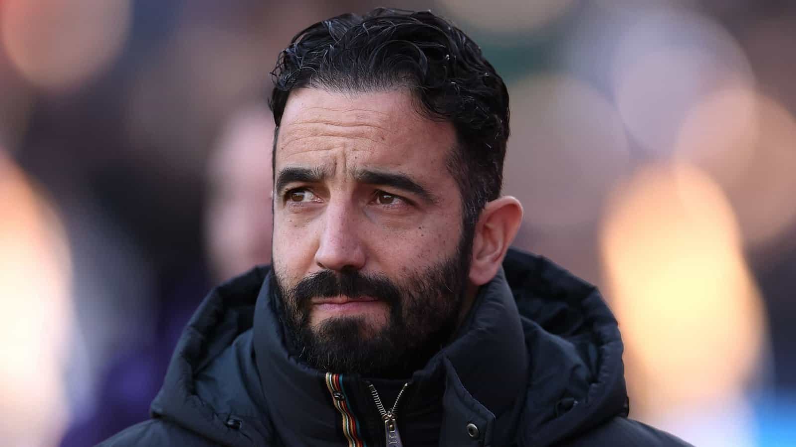 Ruben Amorim, gekleed in een zwarte jas, staart bedachtzaam voor zich, terwijl een moment van intensiteit wordt vastgelegd tegen een wazig stadionachtergrond.