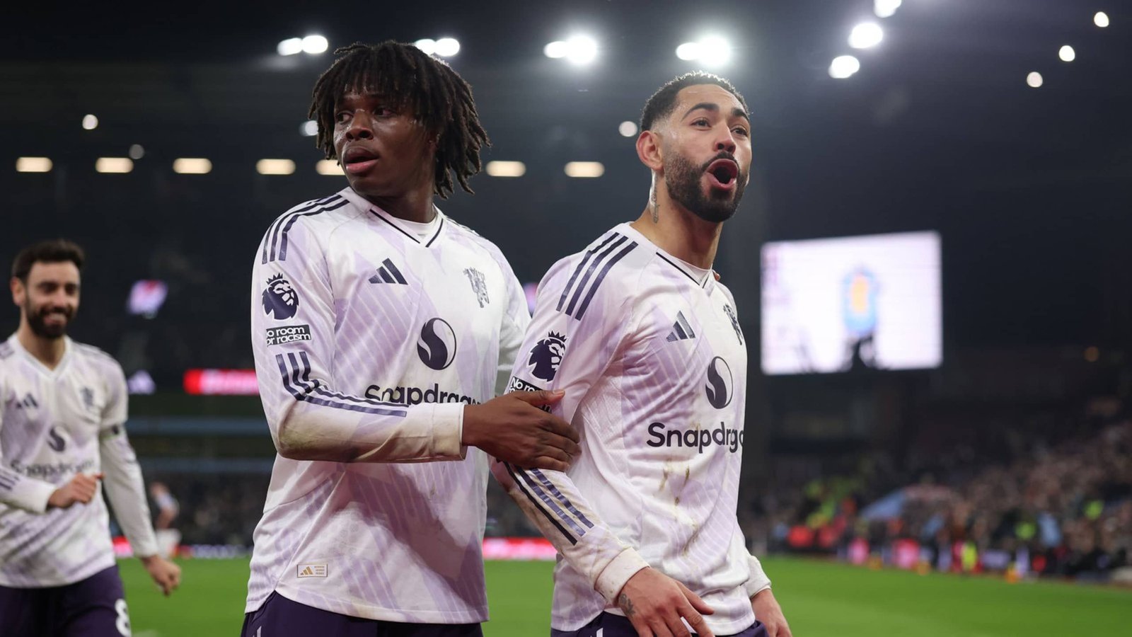 Leeds United-spelers vieren een doelpunt, dragen hun witte uittenue met paarse accenten. Opwinding is zichtbaar op hun gezichten in een vol stadion.