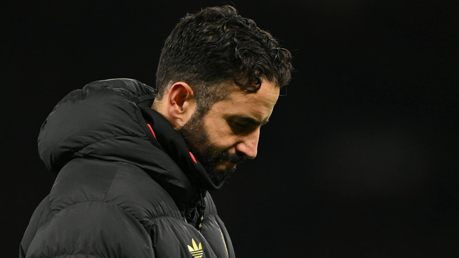 Ruben Amorim, gekleed in een zwarte pufferjack met capuchon, kijkt somber tijdens een gespannen moment, wat zijn problematische positie bij Man Utd benadrukt.