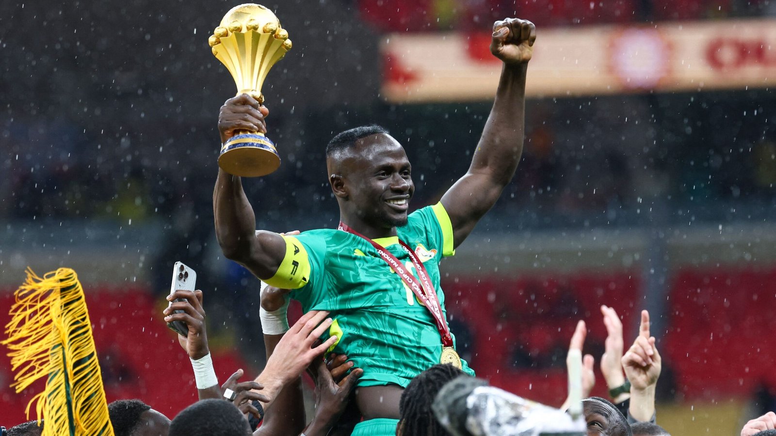 Sadio Mané célèbre triomphalement, brandissant le trophée de la CAN sous la pluie, entouré de coéquipiers en liesse vêtus du maillot vert du Sénégal.