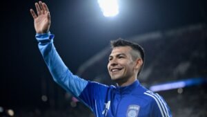 Hirving Lozano in a blue San Diego FC jacket smiles and waves to fans, illuminated by stadium lights during a match.