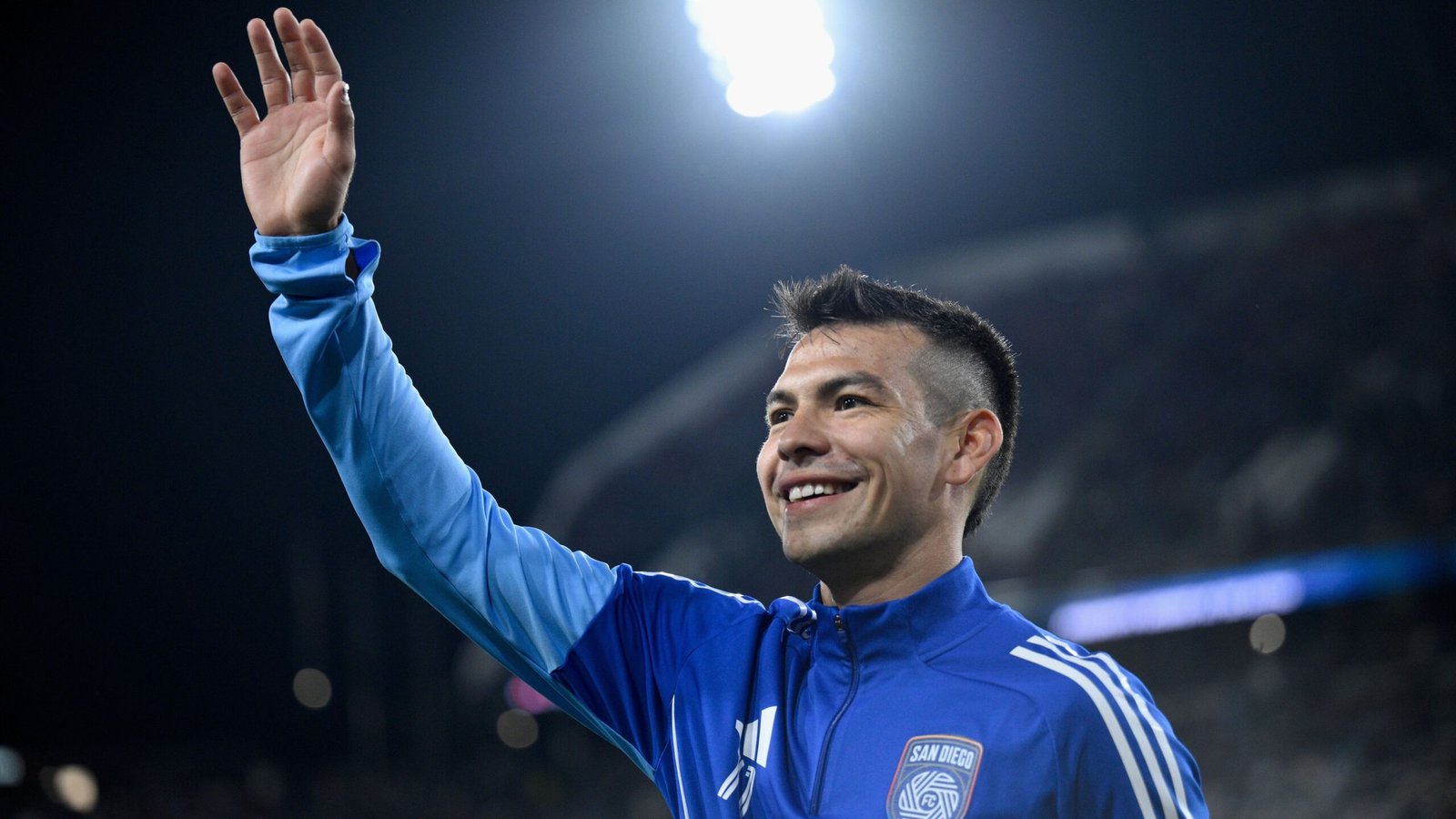 Hirving Lozano in a blue San Diego FC jacket smiles and waves to fans, illuminated by stadium lights during a match.