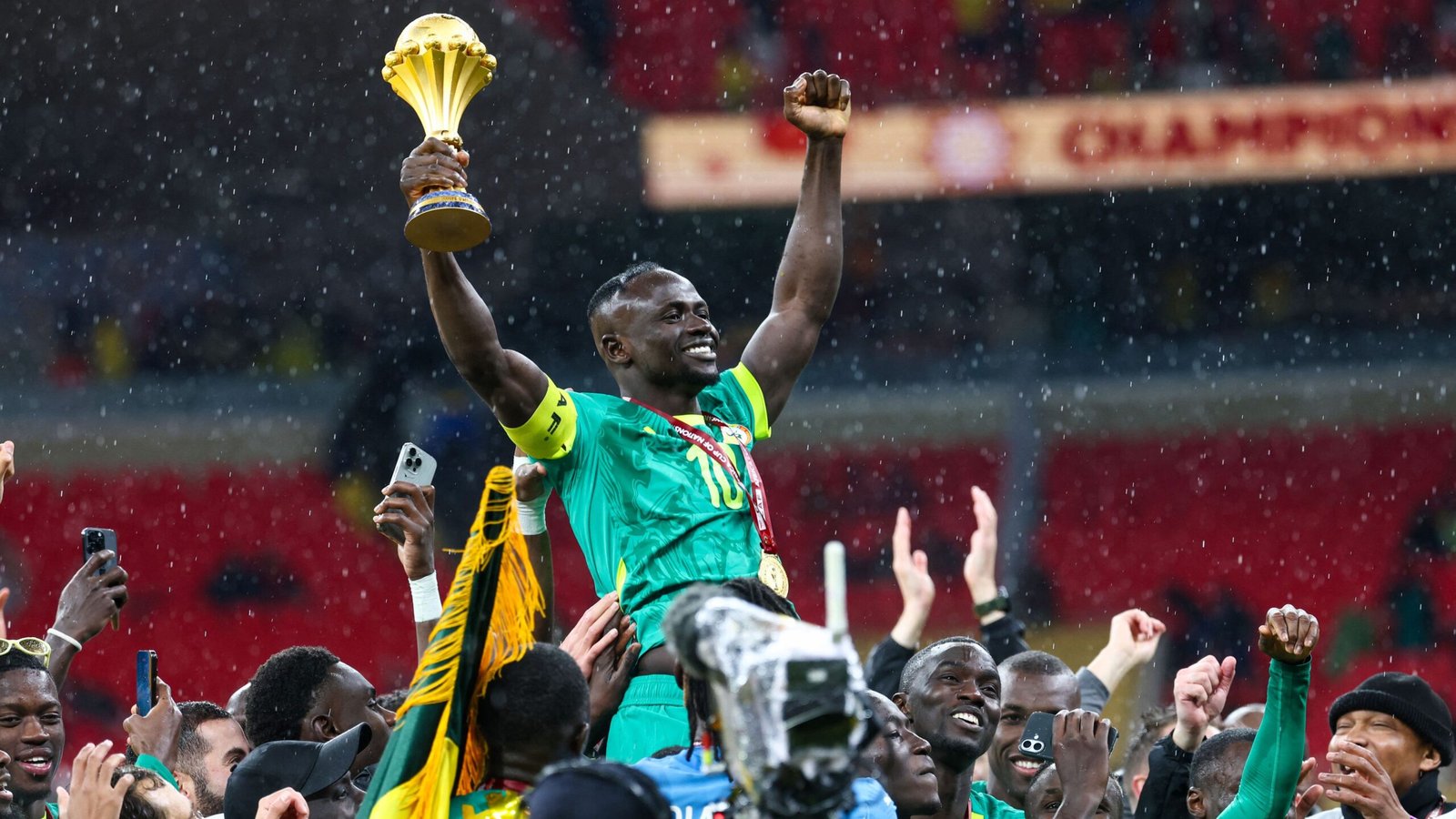 Sadio Mané viert het hijsen van de AFCON-trofee, omringd door teamgenoten in groene shirts, overgoten door regen en vreugde.