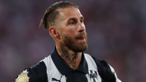 Sergio Ramos portant un maillot de football en noir et blanc, avec des tatouages visibles, l'expression concentrée, debout devant un fond de stade flou.