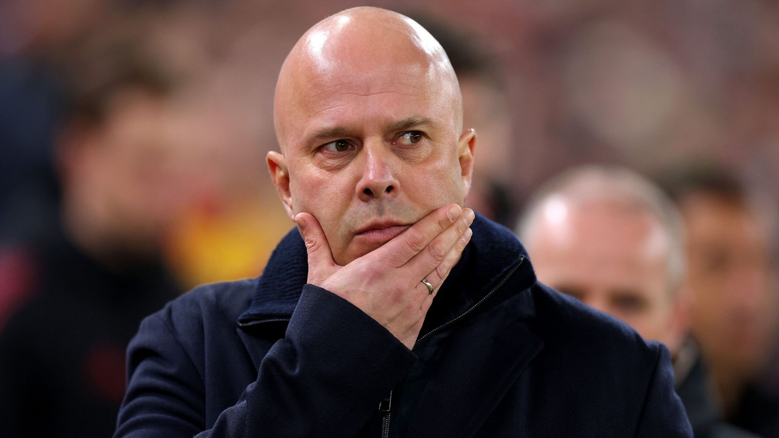 Een bedachtzame coach met een geschoren hoofd, gekleed in een donkere jas, peinst intens tijdens een wedstrijd, met wazige fans op de achtergrond.