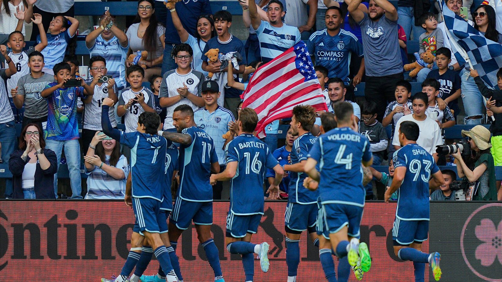 Spelers van Sporting Kansas City vieren een doelpunt, geflankeerd door opgewonden fans in teamtenues, zwaaiend met een Amerikaanse vlag en juichend in een uitverkocht stadion.