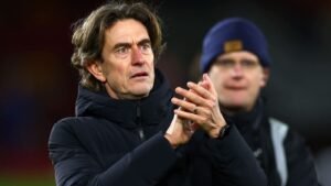 Thomas Frank fait des gestes vers la foule, l'air préoccupé tout en applaudissant. Un membre de son staff se tient derrière lui, portant un bonnet foncé.