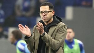 De hoofdcoach van Strasbourg die vanaf de zijlijn applaudisseert tijdens een wedstrijd, gekleed in een nette jas en met een bril, met spelers op de achtergrond.