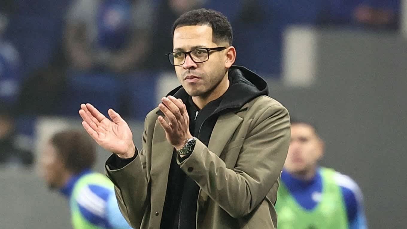 De hoofdcoach van Strasbourg die vanaf de zijlijn applaudisseert tijdens een wedstrijd, gekleed in een nette jas en met een bril, met spelers op de achtergrond.