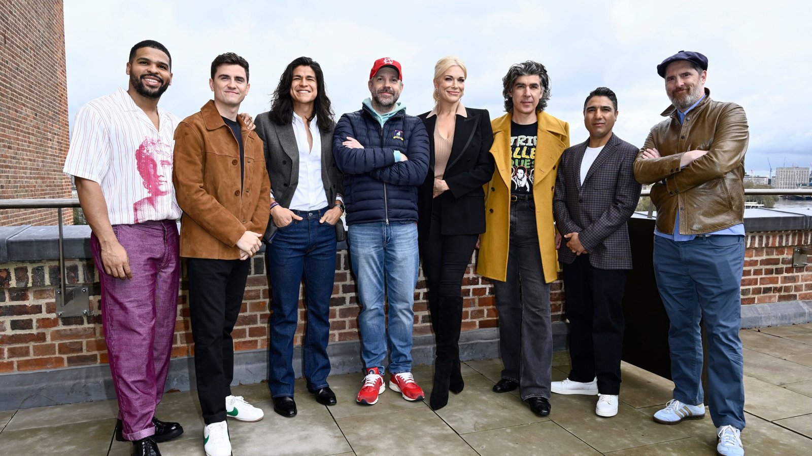Cast of "Ted Lasso" poses on a rooftop, showcasing diverse styles in casual and smart attire, against an urban backdrop.