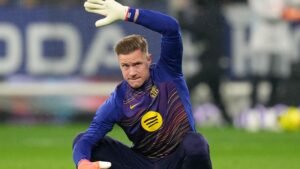 Marc-André ter Stegen in een blauw Barcelona-tenue, zich voorbereidend op het spel met één opgeheven hand, toont zijn geconcentreerde uitdrukking en keepershandschoenen.