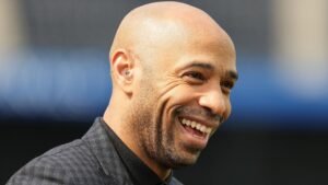 Thierry Henry glimlachend, met zijn kale hoofd en een vrolijke uitdrukking, gekleed in een zwart geruit pak, tegen een wazige achtergrond.