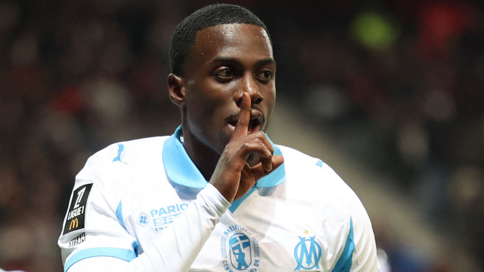 Tim Weah van Marseille gebaart om stilte na het scoren, gekleed in de lichtblauwe en witte outfit van het team, tegen een vervaagde stadionachtergrond.