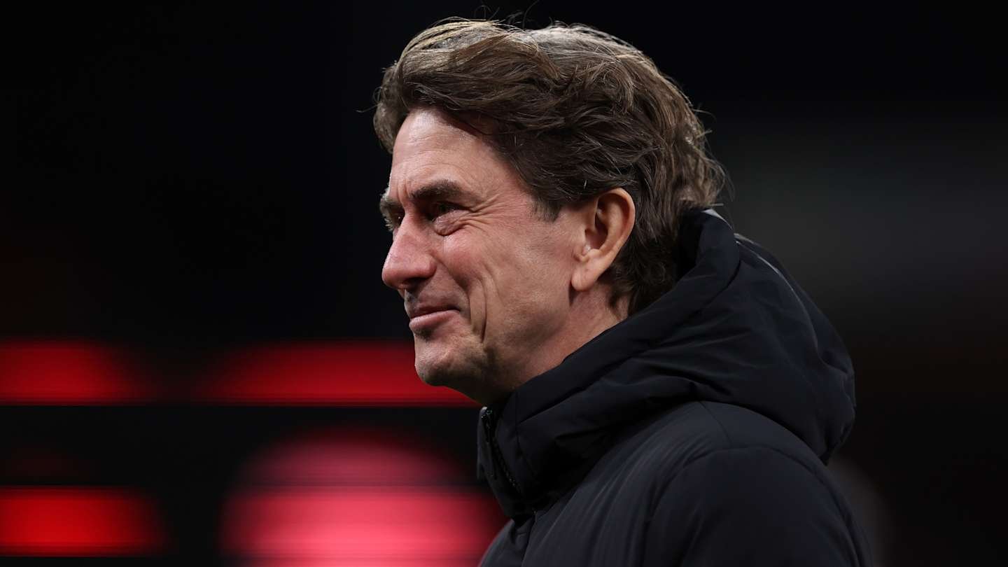 Brentford manager smiles warmly during a match, with a blurred background featuring red lighting and dark tones.