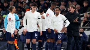 Tottenham-spelers in witte shirts, wachtend op instructies van de coach, met fans op de achtergrond tijdens een wedstrijd.