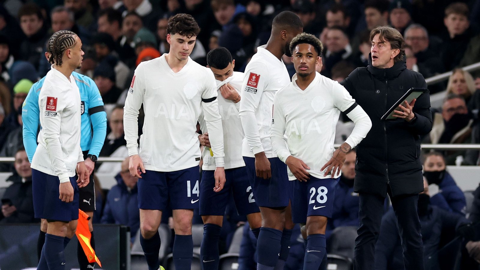 Tottenham-spelers in witte shirts, wachtend op instructies van de coach, met fans op de achtergrond tijdens een wedstrijd.