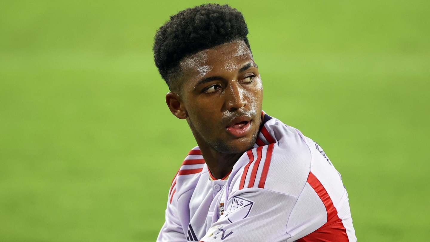 USMNT defender showing concern on the pitch, wearing a white jersey with red accents, against a blurred green backdrop.
