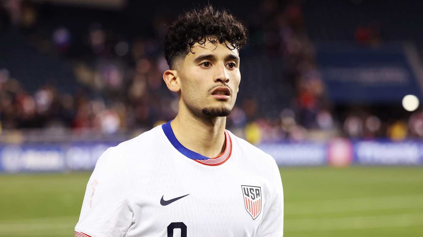 لاعب المنتخب الأمريكي بشعر مجعد، يرتدي قميصًا أبيض مع لمسات من الأزرق والأحمر، يبدو متفكرًا بعد المباراة، مع خلفية ملعب ضبابية.