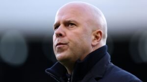 Arne Slot, gekleed in een donkere coltrui, kijkt bedachtzaam in de verte met een serieuze uitdrukking, tegen een vervaagde stadionachtergrond.