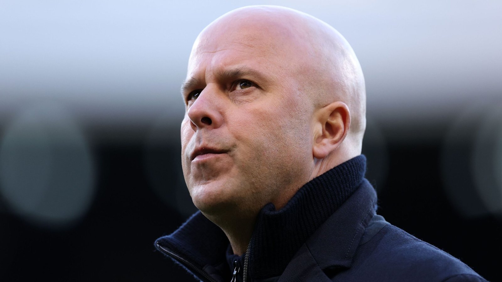 Arne Slot, sporting a dark turtleneck, looks thoughtfully into the distance with a serious expression, set against a blurred stadium background.