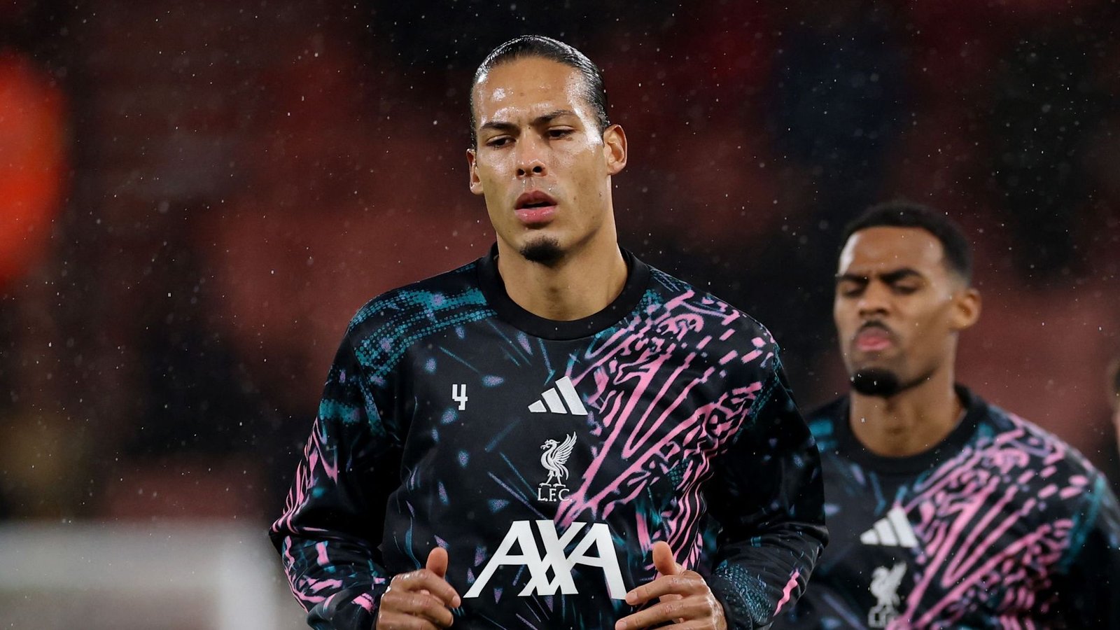 Virgil van Dijk in een opvallend Liverpool-trainingspak, geconcentreerd tijdens een regenachtige warming-up, met een teamgenoot op de achtergrond.