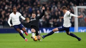 Van de Ven en een andere Spurs-speler dagen een tegenstander in een zwart shirt uit, terwijl ze intense wedstrijdactie vastleggen in het Tottenham Hotspur Stadium.